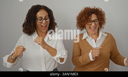 Zwei fröhliche, reife Frauen mit Brille lachen und tanzen zusammen vor einem weißen Wandhintergrund und halten einen lebendigen Moment der Freundschaft und des Glücks fest Stockfoto