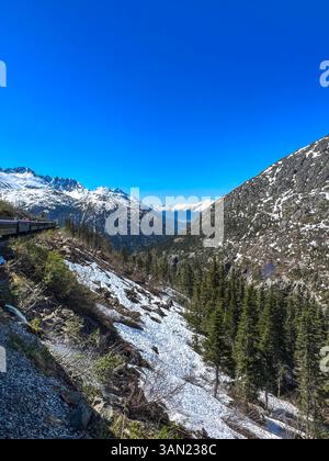 Ein atemberaubender Blick auf einen Zug, der sich durch die zerklüfteten Berge Alaskas schlängelt, vorbei an schneebedeckten Hängen und hoch aufragenden Gipfeln. Der strahlend blaue Himmel verbessert Stockfoto