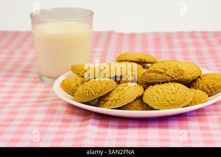 Ein Teller mit Keksen sitzt auf einer rosa-weiß karierten Tischdecke neben einem Glas Milch. Die Kekse sind pyramidenförmig angeordnet, mit der oberen Kappe Stockfoto