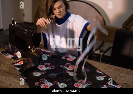Junge Frau Designerin, die schwarzen floralen Stoff auf schwarzer Heimnähmaschine auf Holztisch nähen. Sie hält ein Maßband, fotografiert im Workflow Stockfoto