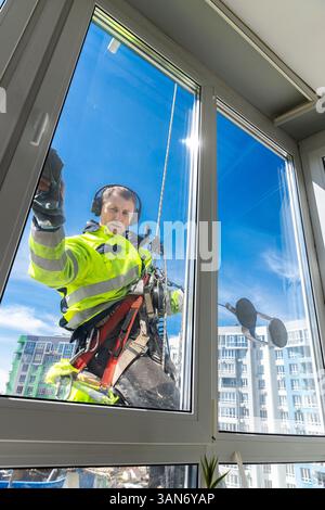 Ein professioneller Fensterreiniger in Sicherheitsausrüstung, der in der Höhe arbeitet, um ein Hochhausfenster mit einem klaren blauen Himmel im Hintergrund zu reinigen Stockfoto