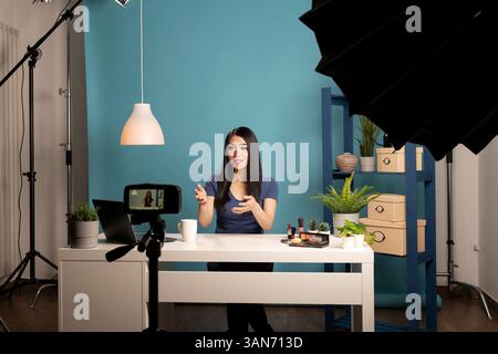 Junger asiatischer Blogger, der ein kosmetisches Rezensionsvideo in einem Studio mit einer Kamera auf einem Stativ aufnimmt. Sie sitzt an einem Schreibtisch mit Make-up-Produkten, mit professioneller Beleuchtung und blauem Hintergrund. Stockfoto