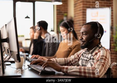 Schwarzer Mann im Rollstuhl und seine multiethnischen Kollegen saßen mit Computern und Headsets an Schreibtischen und kümmerten sich um Kundenanrufe. Männlicher Afro-amerikanischer Kundendienstmitarbeiter, der in einem Arbeitsplatz mit Ziegelwänden arbeitet. Stockfoto