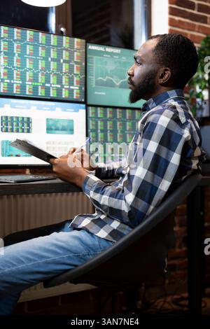 Fokussierter Börsenmakler mit Clipboard, Untersuchung von Forex-Markttrends in der Nacht, Verwendung mehrerer Monitore in modernen Büros. Schwarzer Mann, der Finanzdaten und Aktiendiagramme überprüft, die auf Computerbildschirmen angezeigt werden. Stockfoto