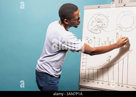 afroamerikanischer männlicher Profi in Freizeitkleidung zeichnet Grafiken und Diagramme auf einem Zeichenbrett. Schwarzer Mann, der neben dem Whiteboard steht und Daten für die Präsentation im Studio mit blauem Hintergrund vorbereitet. Stockfoto