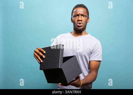 Staunender afroamerikanischer Mann, der eine schwarze Geschenkschachtel öffnet und mit breiten Augen vor blauem Hintergrund reagiert. Schockierte Einzelperson, die ein Geschenk auspackte, staunte über die Lieferüberraschung. Stockfoto