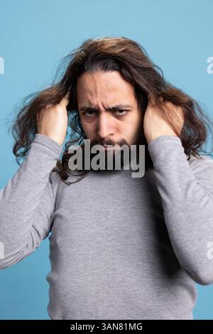 Ein Mann aus dem Nahen Osten verhält sich verzweifelt und zieht sich vor der Kamera die Haare aus, ist wütend und gestresst vor blauem Hintergrund. Junger Erwachsener, der sich überwältigt fühlt und in Verzweiflung, in Not. Stockfoto
