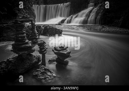 Ein Wasserfall fließt ruhig über Felsen, sorgfältig ausbalancierte Steintürme im Vordergrund, künstlerisches Schwarz-weiß-Bild, Oberallgaeu, Bayern, Deutsch Stockfoto