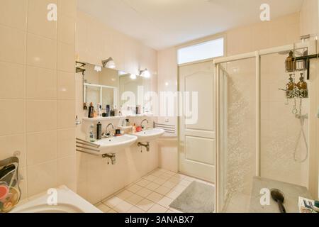 Ein geräumiges und modernes Bad mit zwei Waschbecken, Spiegeln und einer Glasdusche. Die hellen Fliesen und das Tageslicht schaffen eine einladende Atmosphäre. Stockfoto