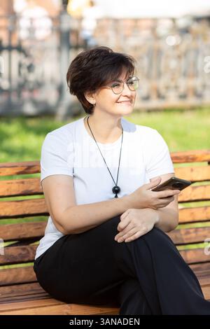 Eine schöne Frau mittleren Alters mit Brille und kurzen dunklen Haaren sitzt auf einer Bank Stockfoto