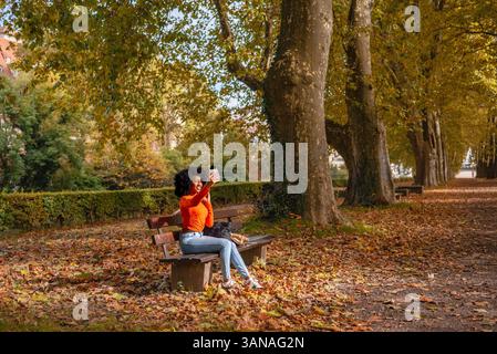 Schöne Frau, die Selfies macht, während sie auf einer Parkbank in einem Herbstummel in Tubingen sitzt. Stockfoto