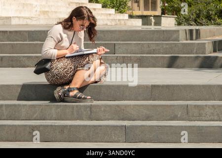 Schöne reife Frau, die auf der Treppe sitzt und ein Buch liest. Stilvolle Frau, die an einem sonnigen Sommertag draußen auf der Treppe ein Buch liest - Tirana Alba Stockfoto