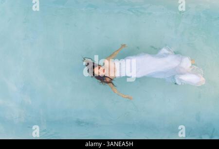 Luftaufnahme über einer attraktiven Frau, die in einem Hochzeitskleid im Wasser schwimmt. Stockfoto