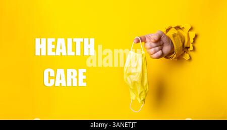 Person, die eine gelbe Maske mit dem Wort Health Care hält, das unten geschrieben ist. Begriff der Vorsicht und Verantwortung gegenüber der Volksgesundheit Stockfoto