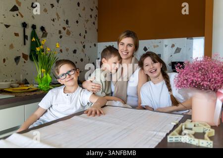 Glückliche Mutter mit Kindern in gemütlicher Küche. Eine lächelnde Mutter umarmt ihre drei Kinder in einer warmen und gemütlichen Küche. Die Familie genießt ein fröhliches und Stockfoto