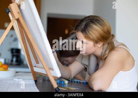 Die Frau konzentrierte sich auf das Malen zu Hause. Eine junge Frau, die zu Hause sorgfältig auf eine Leinwand malt, mit einem Pinsel und Acrylfarben. Stockfoto