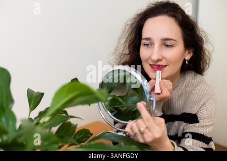 Lächelnde Frau, die Lippenstift mit einem Spiegel und einer Pflanze in der Nähe aufträgt, Eine lächelnde Frau trägt roten Lippenstift auf, mit einem kleinen runden Spiegel, mit einer grünen Pflanze Stockfoto