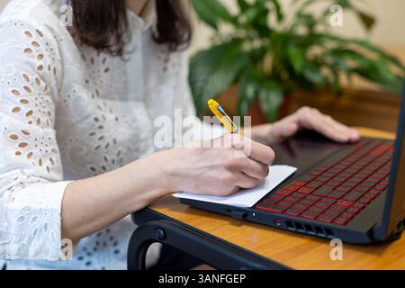 Frau, die Notizen macht, während sie am Laptop arbeitet. Nahaufnahme einer Frau, die in einem Notebook schreibt, während sie einen Laptop benutzt. Stockfoto