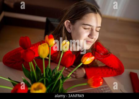 Frau genießt friedlich Tulpen in natürlichem Licht. Eine junge Frau in einem roten Pullover lehnt sich mit geschlossenen Augen über einen leuchtenden Tulpenstrauß Stockfoto