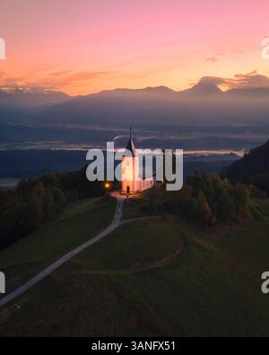 Blick aus der Vogelperspektive auf die St. Primoz Kirche bei Sonnenaufgang auf einem ruhigen Hügel, umgeben von majestätischen Gipfeln der Julischen Alpen, Jamnik, Slowenien. Stockfoto