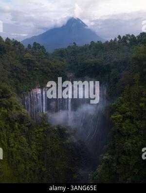 Aus der Vogelperspektive auf einen atemberaubenden Wasserfall mit Rauch, der vom majestätischen Vulkan Merapi, umgeben von üppigem Wald, Tumpak Sewu, Java, Indonesien, aufsteigt. Stockfoto