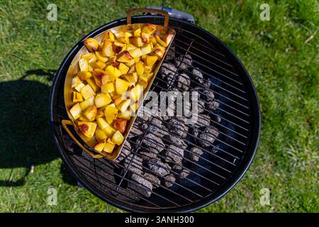 Gegrillte Kartoffeln, die über Holzkohlegrill geröstet werden. Goldbraune Kartoffelstücke, die auf einem Metalltablett auf einem Holzkohlegrill kochen, an einem sonnigen Tag im Freien. Stockfoto