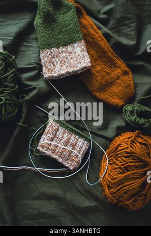 Dunkelgrüner und brauner Garnball mit gestrickten Socken und Stricknadeln. Wollfäden mit tiefgrünem Hintergrund. Gewebte Häkelnaht. Strickhobbyarbeit Stockfoto