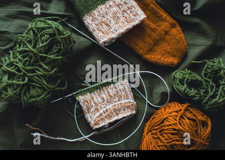 Dunkelgrüner und brauner Garnball mit gestrickten Socken und Stricknadeln. Wollfäden mit tiefgrünem Hintergrund. Gewebte Häkelnaht. Strickhobbyarbeit Stockfoto