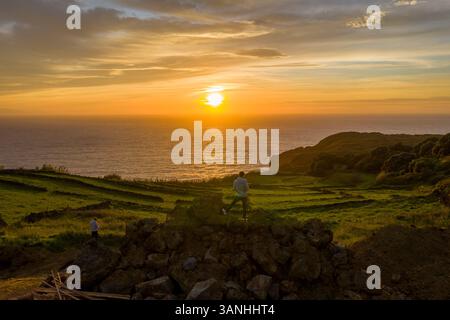 Aus der Vogelperspektive einer Person, die auf dem Felsen steht und den Sonnenuntergang über dem Meer auf der Insel Terceira, Azoren, Portugal, sieht. Stockfoto