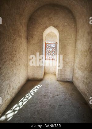 Ein schmaler, alter Steinkorridor mit einem dekorativen Gitterfenster, das komplizierte Schatten auf den Boden wirft. Stockfoto