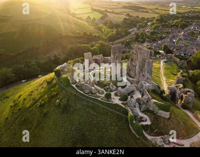 Blick aus der Vogelperspektive auf die historischen Ruinen von Corfe Castle, umgeben von einem malerischen Dorf und grünen Hügeln bei Sonnenuntergang, Corfe Castle, England. Stockfoto
