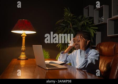 Junge schwarze Geschäftsfrau, die schwere Kopfschmerzen hat, während sie nachts in ihrem Büro mit Laptop arbeitet, ihre Schläfen massiert und unter Augenanstrengung leidet Stockfoto