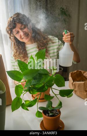 Die lächelnde Frau mit lockigen Haaren, die legere Kleidung und Schürze trägt, sprüht Wasser auf die Zimmerpflanze im Terrakotta-Topf, der zu Hause neben dem Fenster steht und ihr Gartenhobbjekt genießt Stockfoto