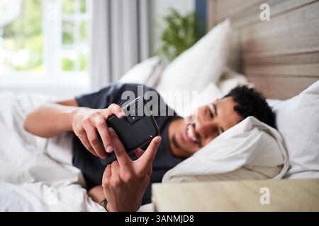 Junger Mann, der sich im Bett entspannt, während er das Smartphone im hellen Zimmer benutzt Stockfoto