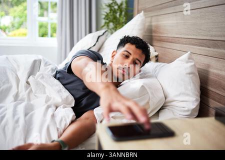 Junger Mann, der im Bett liegt und in einem hellen, modernen Schlafzimmer nach Smartphone greift Stockfoto