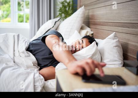 Junger Mann, der nach Wecker greift, während er im Bett in gemütlicher Schlafzimmerumgebung liegt Stockfoto