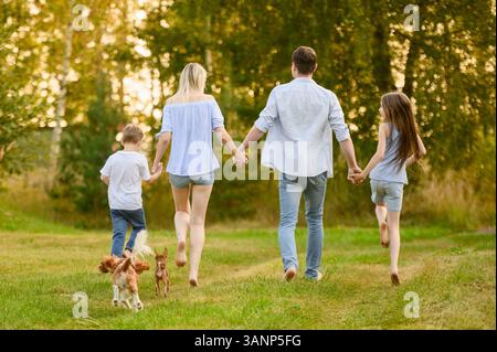 Familie läuft barfuß mit Händen auf Gras, Blick von hinten, zusammen mit Hund. Mom, Dad, Tochter und Sohn genießen Urlaub auf dem Land, laufen und Spaß miteinander haben. Sommerwochenende-Konzept Stockfoto