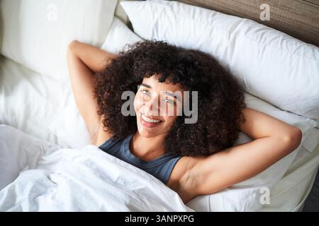 Junge Frau mit natürlichen lockigen Haaren, die sich im Bett entspannen und lächeln Stockfoto