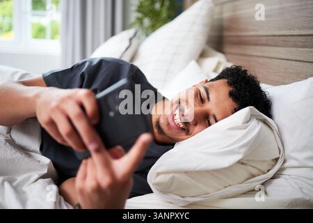Junger Mann, der sich im Bett entspannt, lächelt, während er sein Smartphone drinnen benutzt Stockfoto