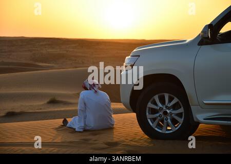 Rub Al Khali Wüste, Sultanat von Oman. Ortsansässiger omanischer Mann in traditioneller Kleidung, der in der Nähe eines Geländewagens auf einer Sanddüne in der Wahiba Sands Dese sitzt Stockfoto