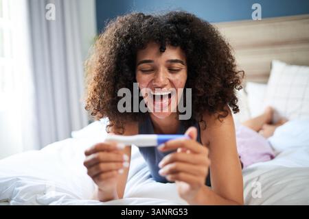 Begeisterte junge Frau mit natürlichem Haar, die auf positiven Schwangerschaftstest im gemütlichen Schlafzimmer reagiert Stockfoto