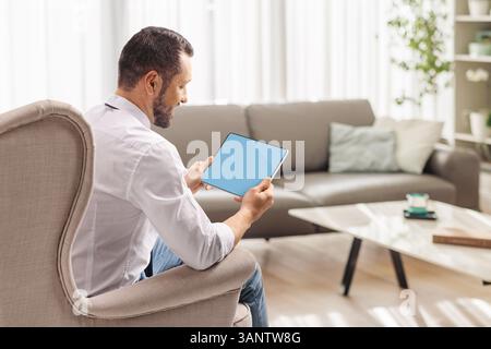 Mann, der zu Hause in einem Sessel sitzt und ein digitales Tablet in einem Wohnzimmer verwendet Stockfoto