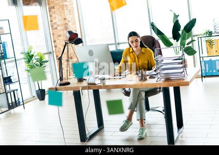 Junge Geschäftsfrau, die in einem stilvollen Büroumfeld arbeitet, umgeben von Postnotizen, Stapeln von Dokumenten und moderner Inneneinrichtung Stockfoto