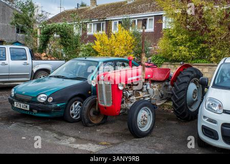 Ein alter roter Traktor parkte neben Autos vor einer Reihe von Häusern im Stadtzentrum von Bridport, Dorset, England. Schrullig. Städtisch-Ländlich. Landleben. Stockfoto