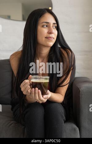 Eine fröhliche Frau sitzt mit einer Kaffeetasse in der Hand auf dem Sofa und lächelt sanft, während sie geduldig in einer gemütlichen und eleganten Umgebung wartet. Stockfoto