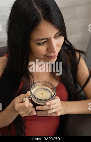 Von oben hält eine Frau mit beiden Händen eine Kaffeetasse, während sie friedlich sitzt und einen warmen und introspektiven Moment zu Hause festnimmt. Stockfoto