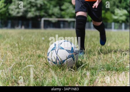 Nahaufnahme der Beine von Kindern in schwarzen Shorts und Socken, die gerade den Fußball auf dem Grasfeld spielen. Heller sonniger Tag, Torpfosten und Bäume im Hintergrund, Akti Stockfoto