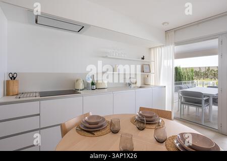 Saubere, moderne weiße Küche mit minimalistischem Design und Glastüren, die zu Sitzgelegenheiten im Freien führen. Stockfoto