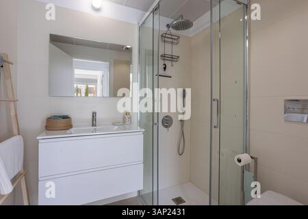 Modernes Bad mit bodenebener Glasdusche, großem Spiegel und sauberem weißen Kosmetikbereich. Stockfoto