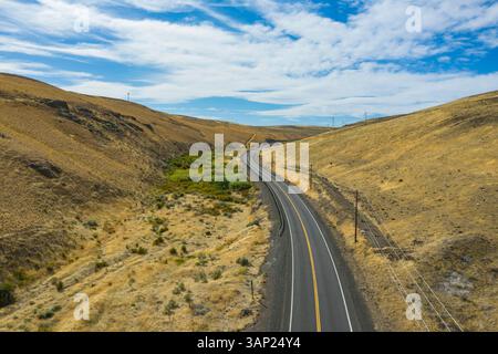 Aus der Vogelperspektive auf der zweispurigen Straße, die sich durch eine ruhige Landschaft und Hügel unter einem sonnigen Himmel mit Wolken schlängelt, Kittitas County, USA. Stockfoto